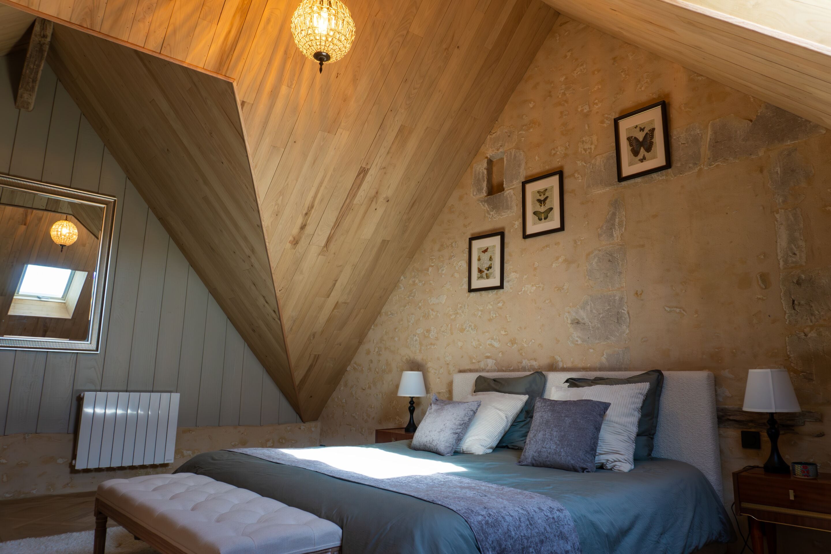 A bedroom with a sloped ceiling, a bed with multiple pillows, a bedside table with a lamp, and framed pictures on the wall.