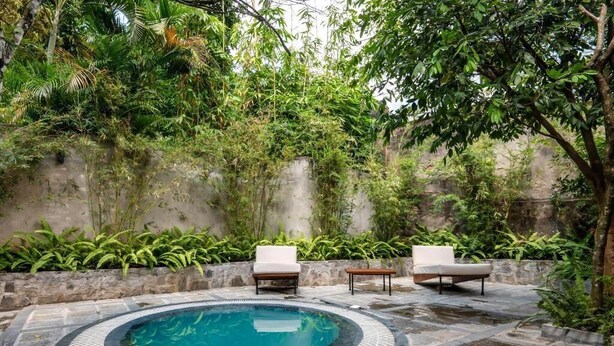Uma piscina circular com borda azulejada, rodeada por piso de pedra, com duas cadeiras de descanso brancas e uma pequena mesa.