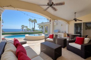 Un salon avec coin détente au bord de la piscine, doté de sièges extérieurs, d’un jacuzzi et d’un espace barbecue, offrant une vue imprenable sur un ciel bleu azur et l’océan.
