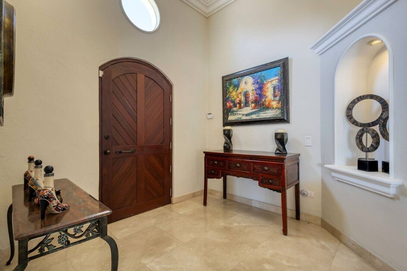 A hallway with a wooden door, a desk with drawers, a painting on the wall, and a decorative shelf with rings.
