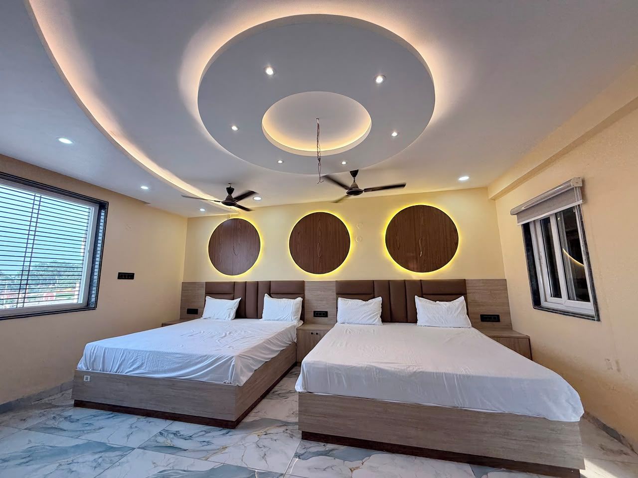 A hotel room with two beds, a circular ceiling design, ceiling fans, and a window with blinds.
