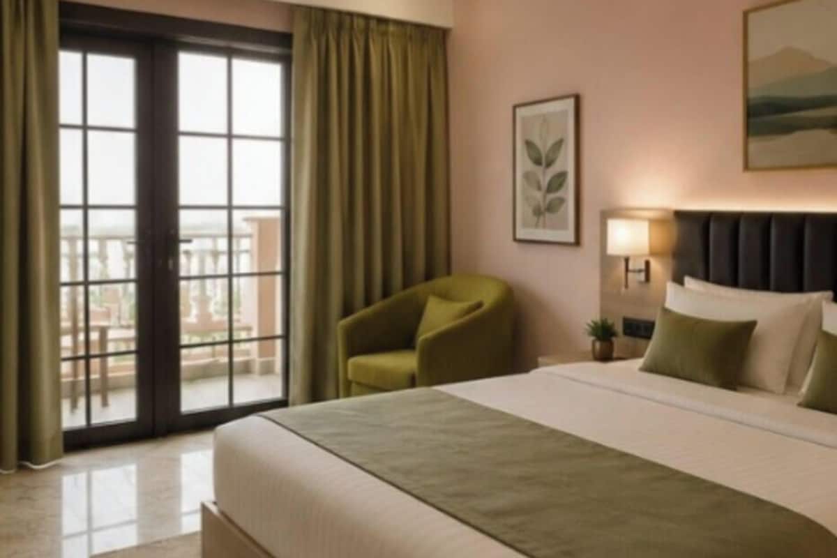 A hotel room with a bed, a chair, a window with curtains, a framed picture, and a lamp.