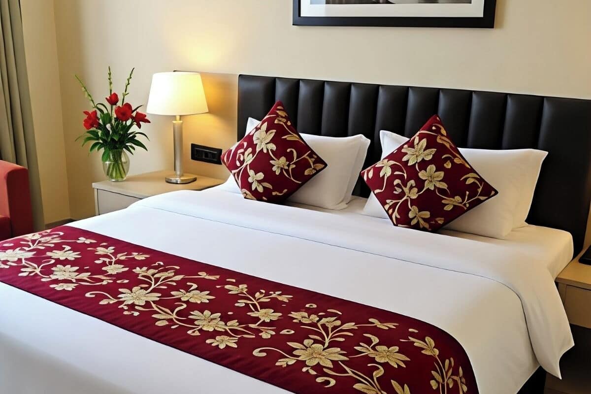 Una habitación de hotel con una cama bien hecha, un cuadro en la pared, un florero con flores y una lámpara en la mesita de noche.
