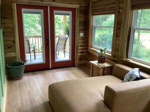 Et værelse med trægulv, en beige sofa, et lille bord med en potteplante og en skydedør, der fører til et udendørs område med en stol og et bord.