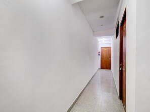 A corridor with white walls, a wooden door at the end, and a tiled floor. - Super Collection O Sector 18 (Noida)