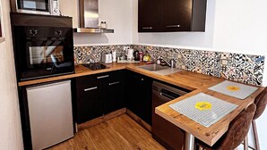 A compact kitchen with a microwave, oven, and dishwasher, a wooden dining table with two chairs, and a patterned backsplash.