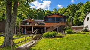 A two-story wooden house with a balcony, a covered porch, and a stone staircase leading to the entrance. - Stillwater on Doe - Private Lakefront Escape (Camdenton)