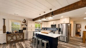 A modern kitchen with wooden flooring, a central island with stools, stainless steel appliances, and a rustic ceiling beam. - Stillwater on Doe - Private Lakefront Escape (Camdenton)