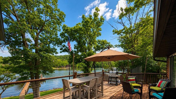A wooden deck with a table and chairs, a grill, and an umbrella. - Stillwater on Doe - Private Lakefront Escape (Camdenton)
