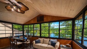 A room with a wooden ceiling, large windows, a sofa, a dining table with chairs, and a ceiling fan. - Stillwater on Doe - Private Lakefront Escape (Camdenton)