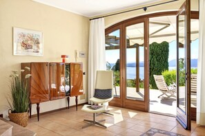 Ein Zimmer mit Holzschrank, einem Sessel, einem Blumentopf und Blick durch eine Glastür auf einen Außenbereich mit Stuhl und Grünpflanzen.