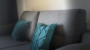Harmaa sohva, jossa on turkoosinsiniset koristetyynyt, seinällä oleva taulu ja ikkuna, jossa on verhot.