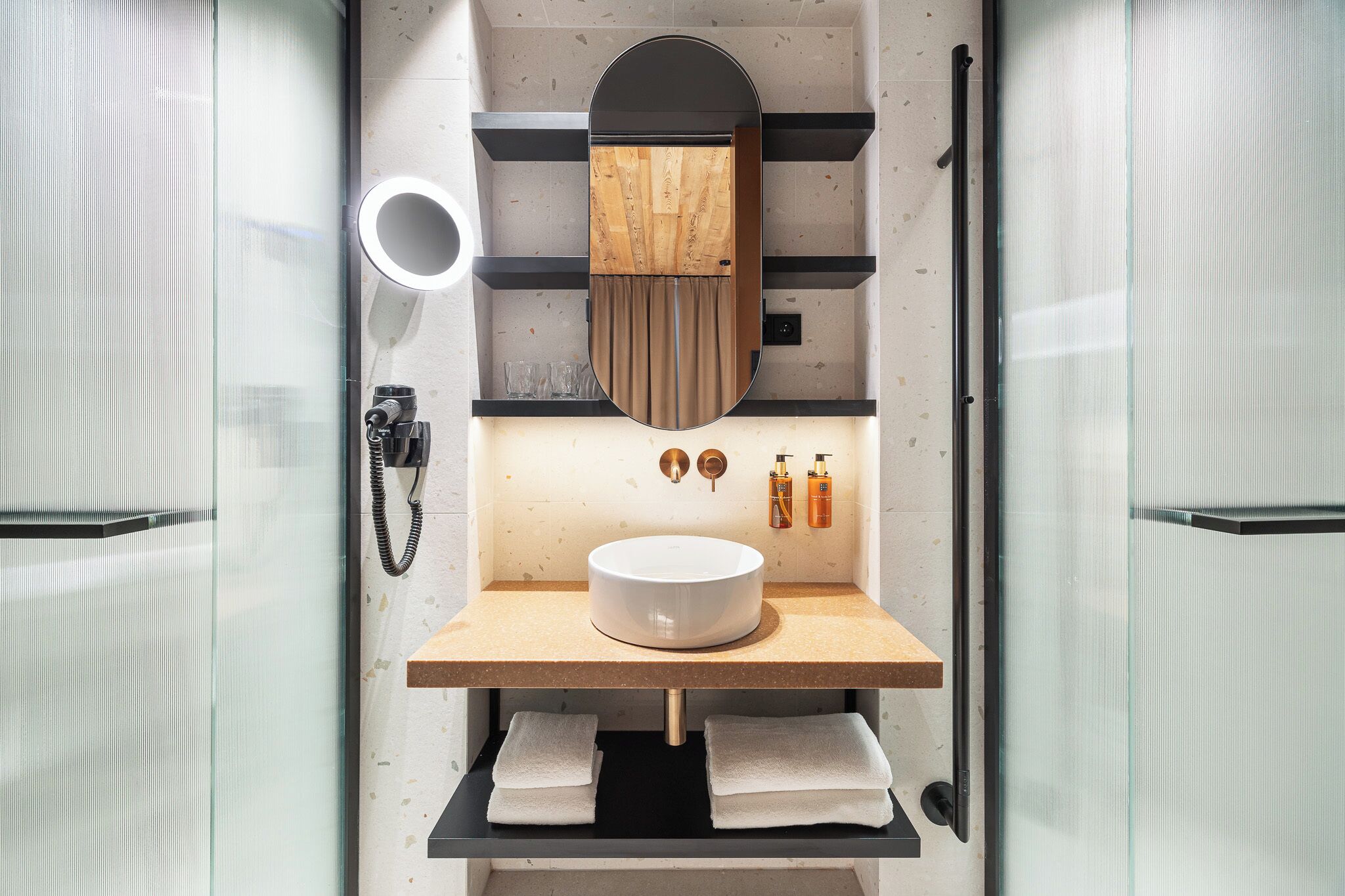 Un baño moderno con cabina de vidrio, tocador de madera, espejo redondo, lavamanos y dos barras para toallas.