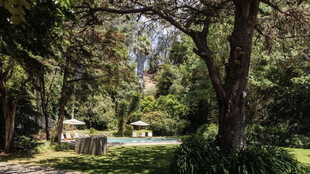 Uma piscina rodeada por vegetação exuberante, um caminho de pedra e um gazebo com guarda-sóis.