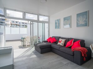 Ein modernes Wohnzimmer mit einem grauen Ecksofa, roten Kissen und einer Glastür zu einem Außenbereich.