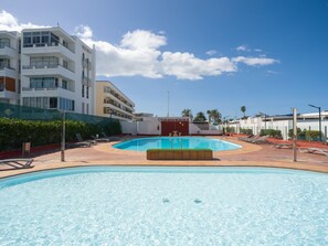Ein Swimmingpool mit Liegestühlen, ein Gebäude mit mehreren Balkonen und im Hintergrund Palmen.
