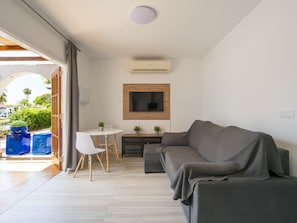 Ein modernes Wohnzimmer mit einer Couch, einem kleinen Tisch und Stühlen. An der Wand ist ein Fernseher angebracht, darüber befindet sich eine Klimaanlage.