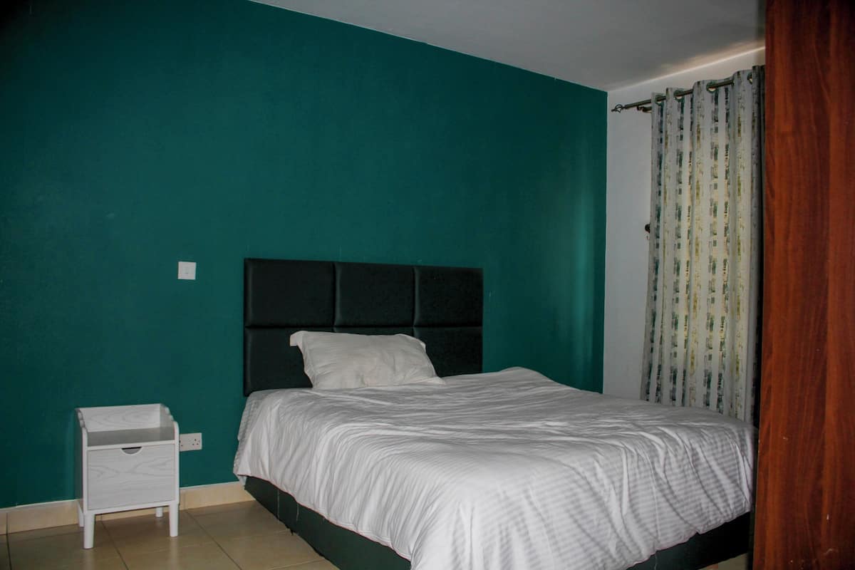 Un dormitorio con cama, mesita de noche, paredes color verde azulado y cortina.