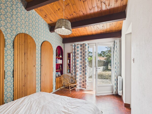 Ein Schlafzimmer mit einem Bett, einem Holzschrank, einem Korbstuhl, einem Bücherregal und einer Schiebetür zum Außenbereich.
