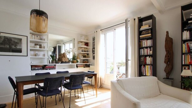 Un salon avec un canapé blanc, une table à manger avec des chaises, une bibliothèque et une grande fenêtre.