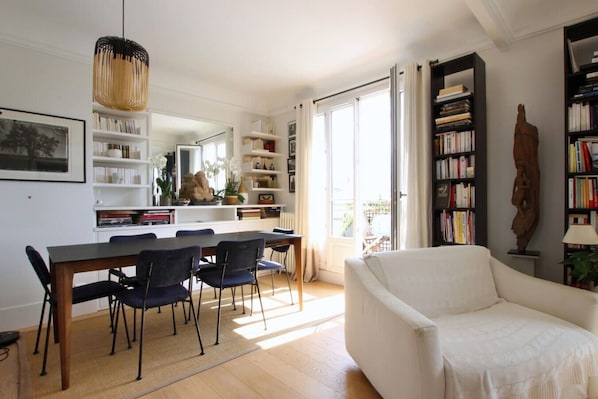 A living room with a white sofa, a dining table with chairs, a bookshelf, and a large window. - Stunning flat with terrasse - Trocadéro (Paris)