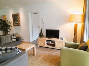 Ein Wohnzimmer mit einer Couch, einem Couchtisch, einem Fernseher auf einem Holzboden, einem kleinen Weihnachtsbaum und einer Lampe.
