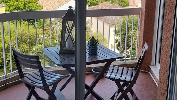 Un balcón con mesa y sillas, una planta en maceta y vistas al mar y al verdor.