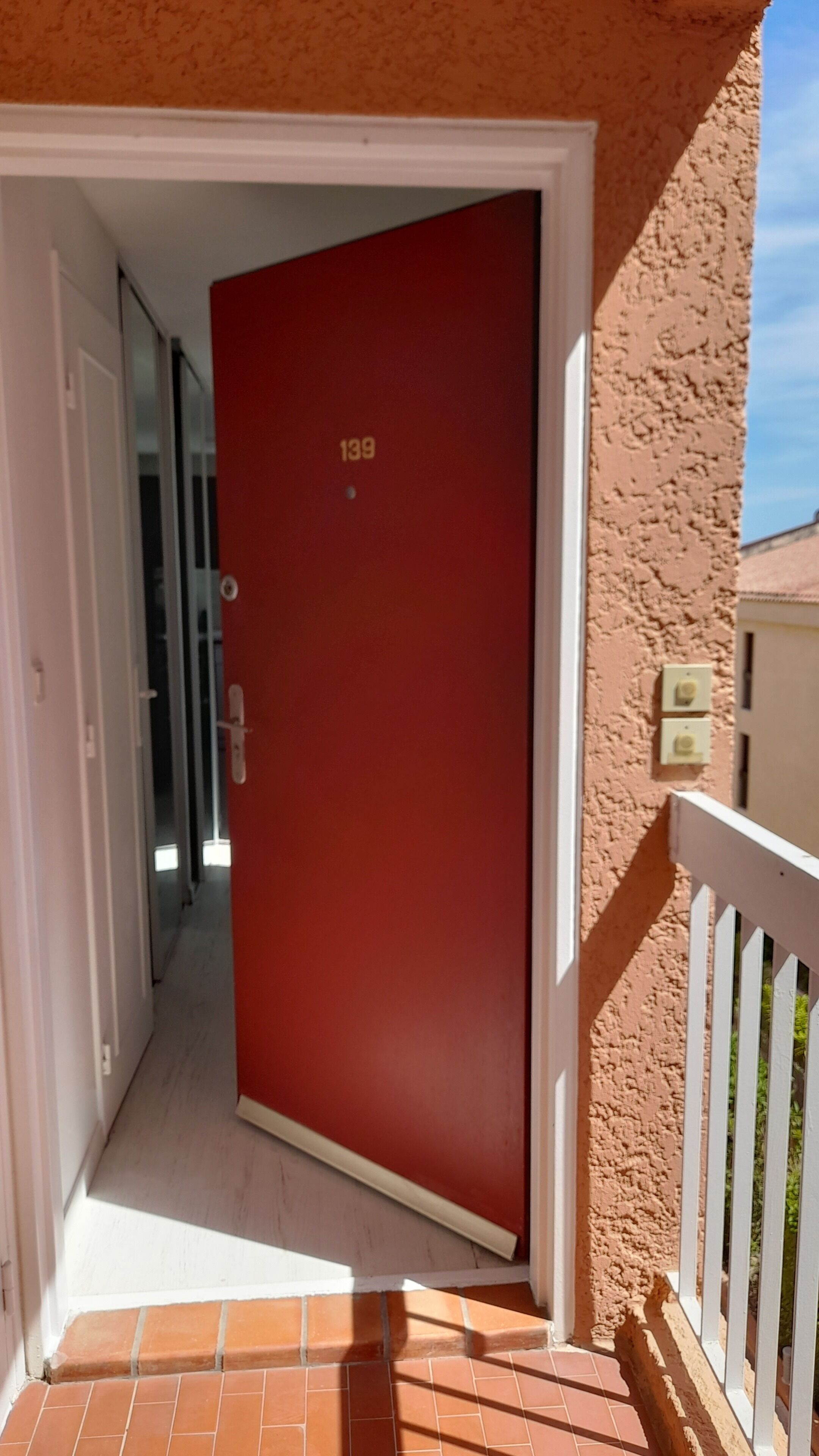 A red door with the number 139, a white door frame, a white railing, and a brick floor.