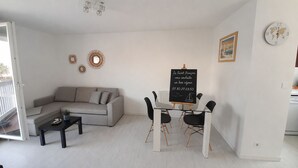 Una sala de estar con un sofá, una mesa de centro, una mesa de comedor con sillas, una pizarra con texto, espejos decorativos y un reloj en la pared.