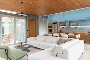 A modern living room with a white sofa, a green armchair, and a coffee table. There is a kitchen area with a refrigerator, microwave, and wooden cabinets.