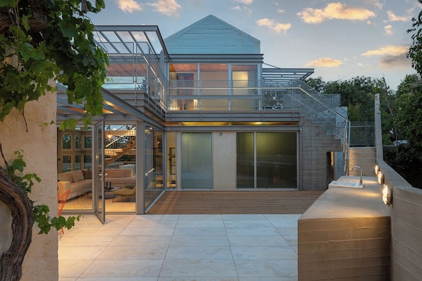 A modern house with a glass facade, a wooden deck, and a staircase leading to a second floor.