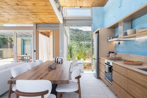 A modern kitchen with a wooden table and white chairs, a built-in oven, and a view of greenery outside. - Anassa House with Rooftop Jacuzzi (Agios Pavlos)