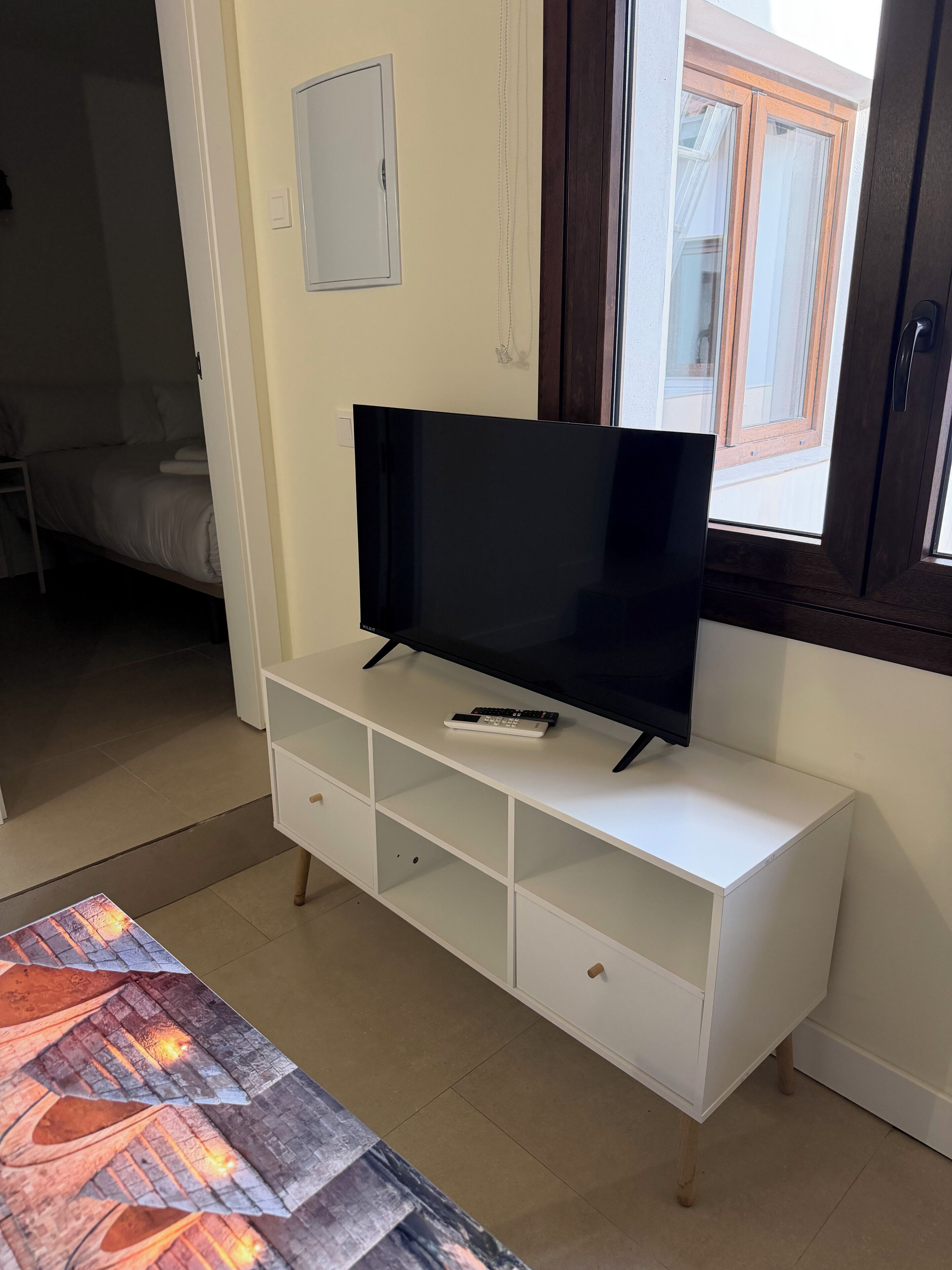 Une pièce avec un téléviseur posé sur un meuble blanc, un lit à l’arrière-plan et une fenêtre avec des rideaux.