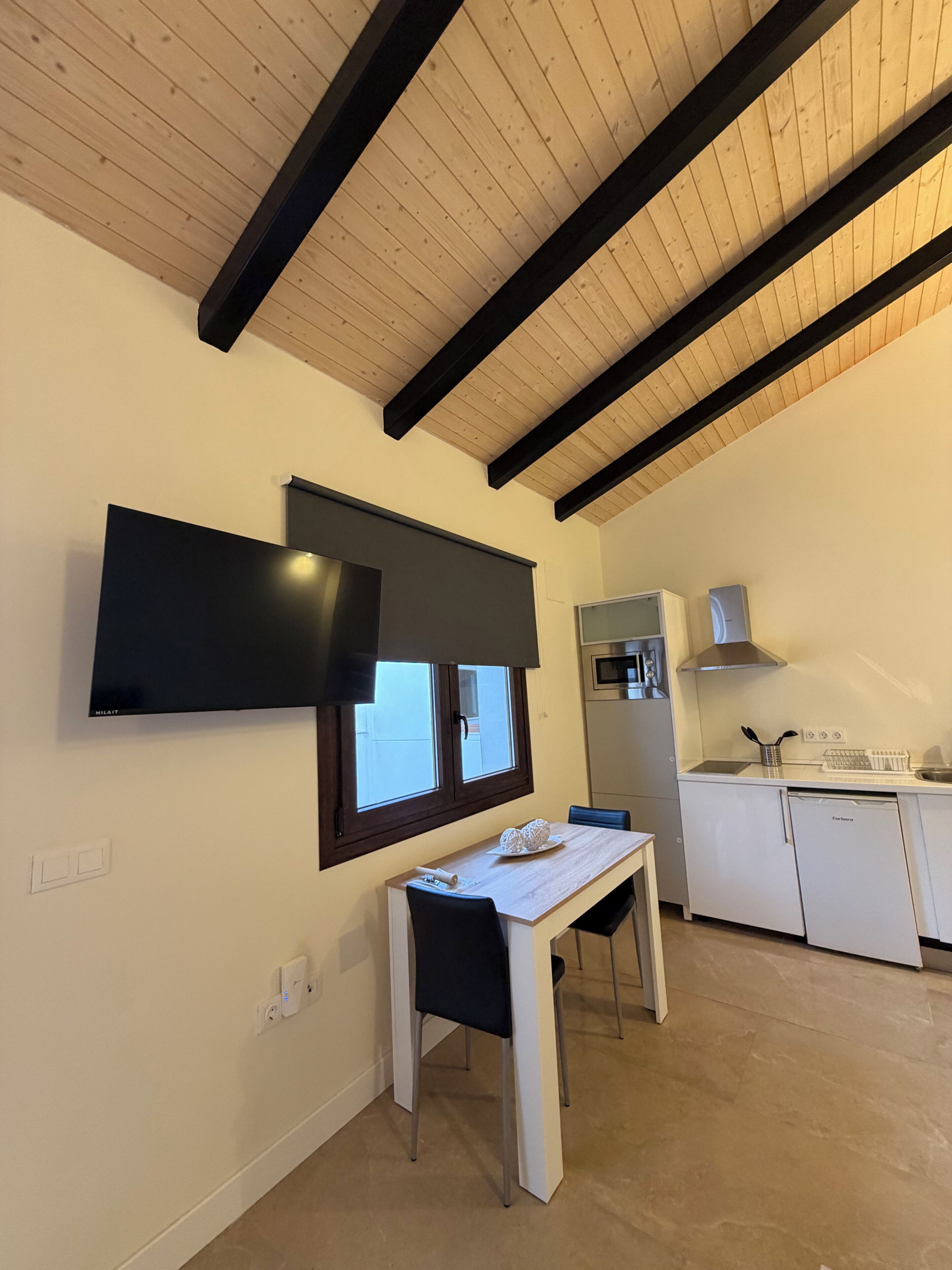A modern kitchen with a built-in oven, a white refrigerator, and a microwave. There is a dining area with a table and chairs, and a flat-screen TV mounted on the wall.