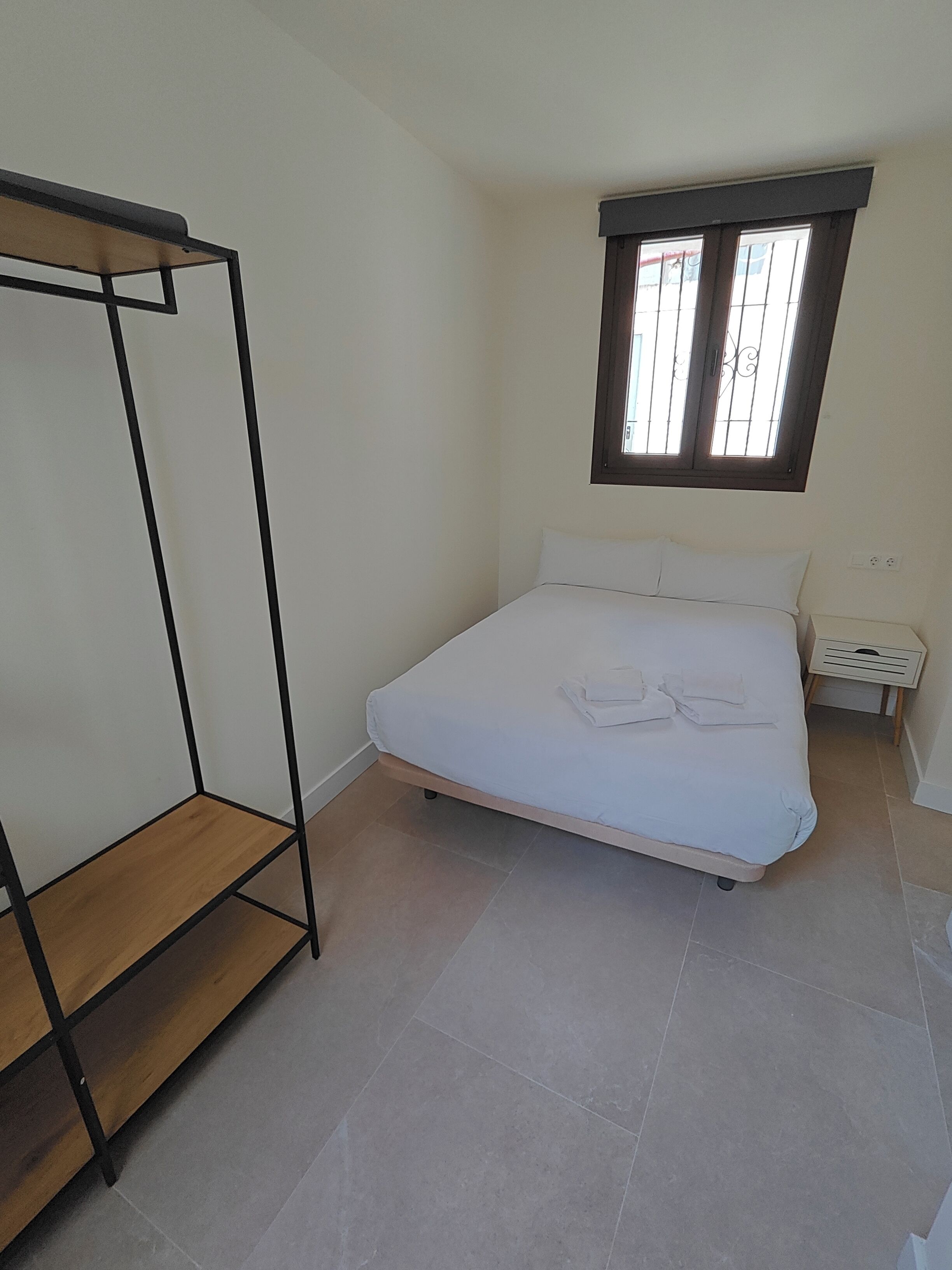 Une chambre à coucher avec un lit, une étagère en bois, une fenêtre avec des barreaux et une table de chevet.