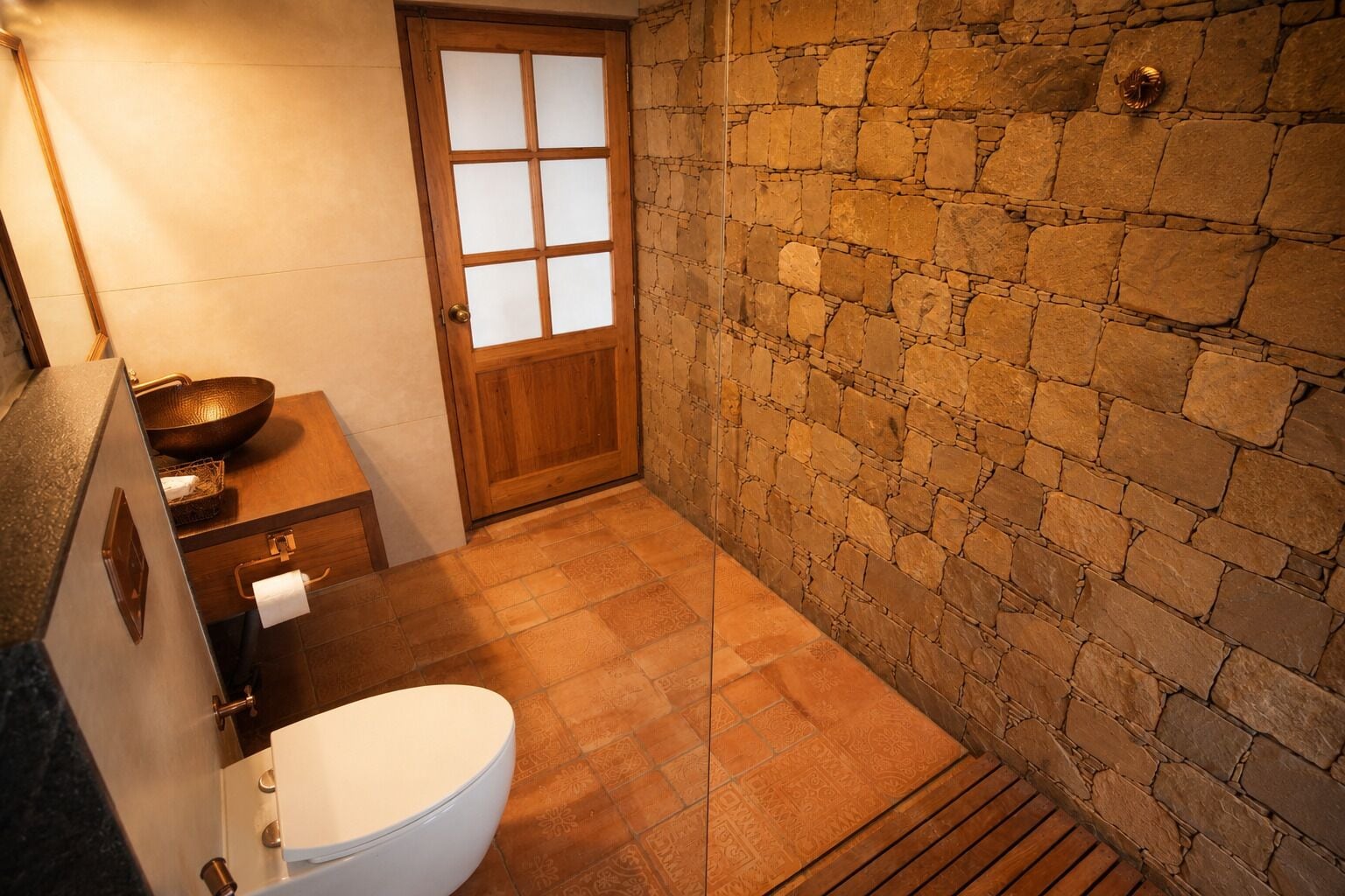 Un baño con pared de piedra, puerta de madera, inodoro blanco y lavabo.