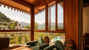 A wooden interior with a sofa and a table, large windows with decorative trim, and a view of a mountainous landscape. - The Lake Heritage Naukuchiyatal (Bhimtal)