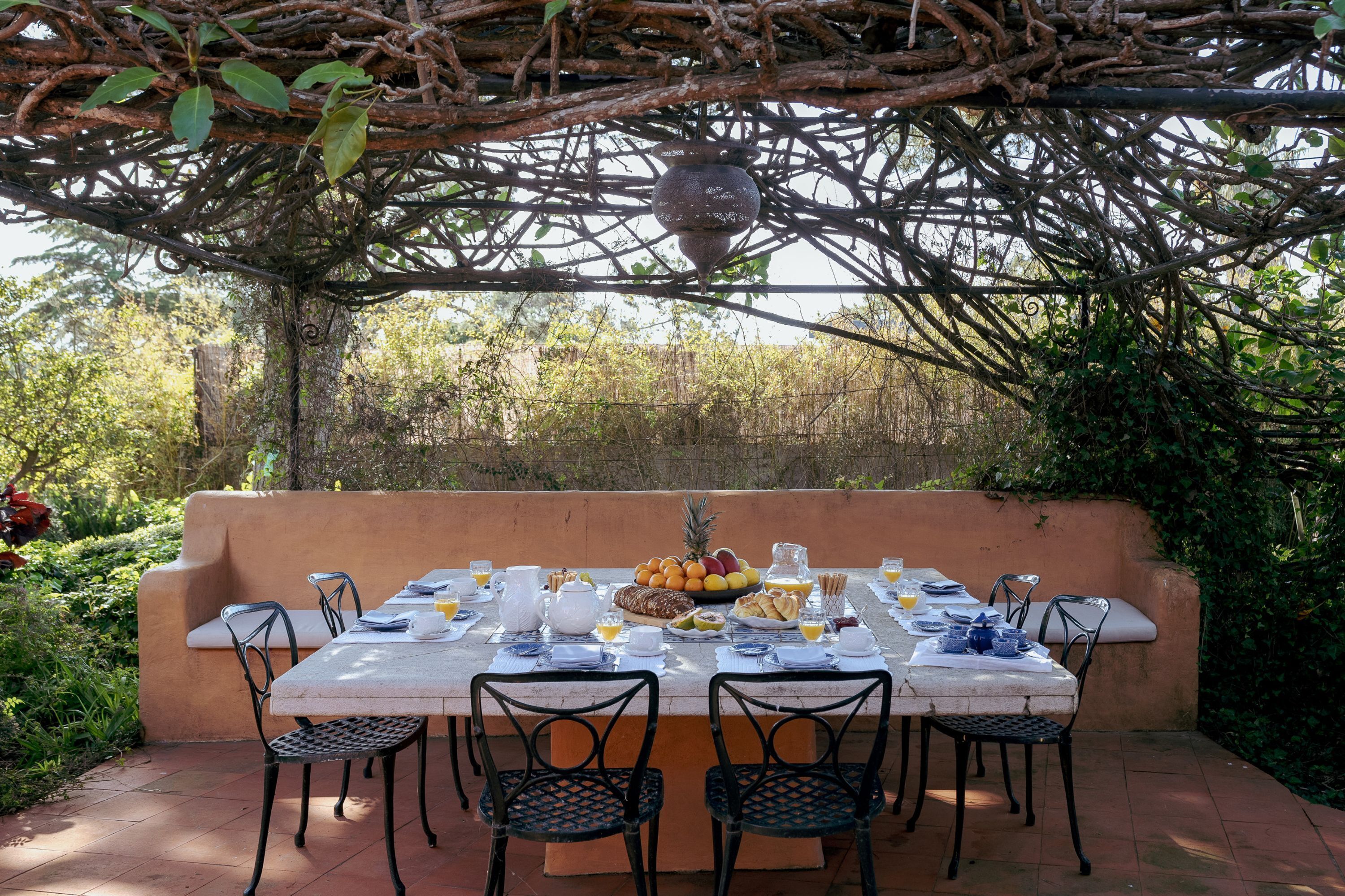 An outdoor dining area with a table set for four, surrounded by chairs, under a natural canopy.