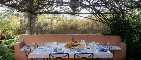 An outdoor dining area with a table set for four, surrounded by chairs, under a natural canopy.