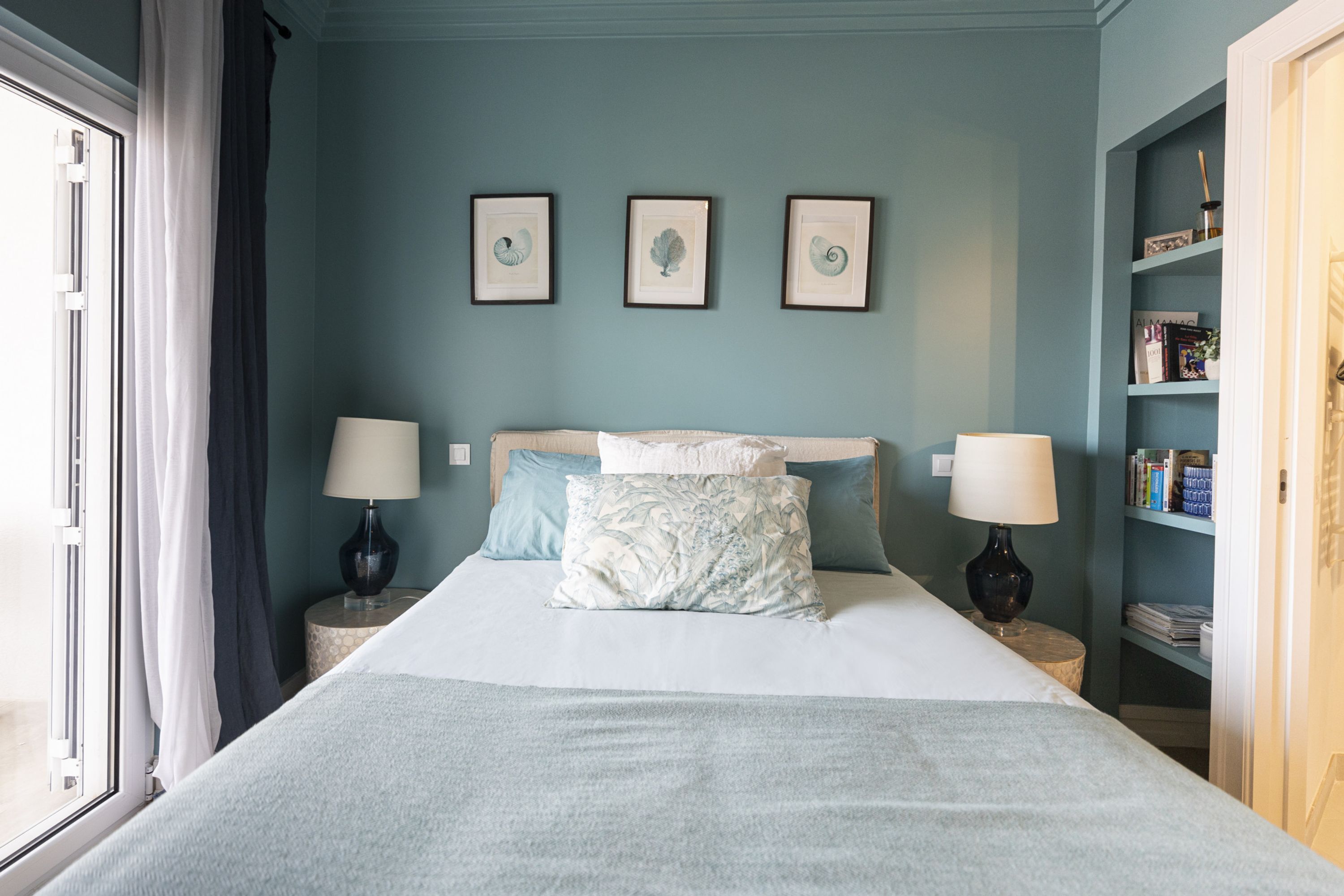 A bedroom with a bed, two bedside tables with lamps, a window with curtains, and a bookshelf with books.
