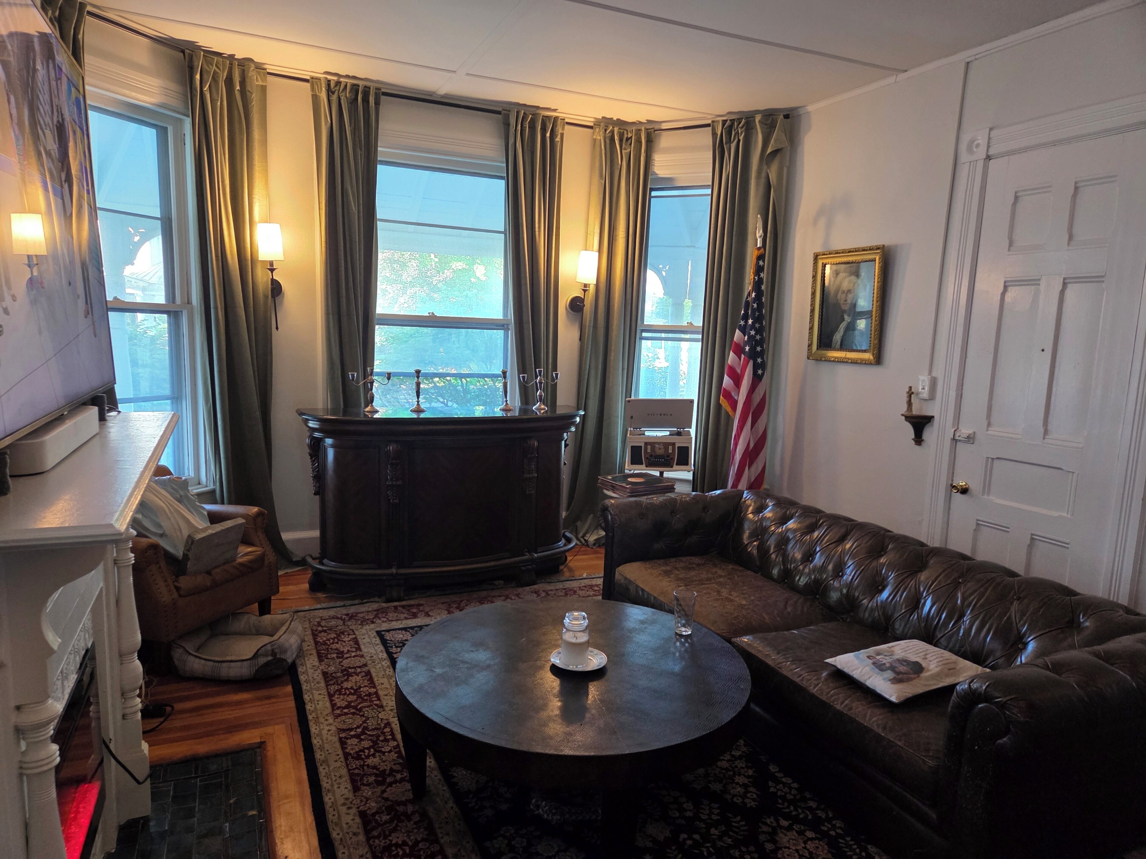 Uma sala de estar com sofá de couro marrom escuro, mesa de centro redonda, lareira, janela grande com cortinas e uma bandeira americana.