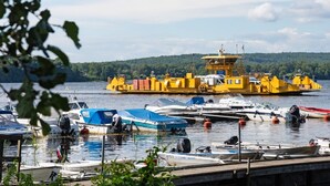 Un muelle de transbordador con varios barcos, incluyendo un gran transbordador amarillo, y una ribera boscosa al fondo.
