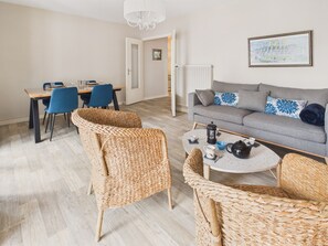 Ein modernes Wohnzimmer mit grauem Sofa, Korbstühlen, einem Esstisch mit Stühlen, einem Couchtisch mit Teekanne und einer Heizung.