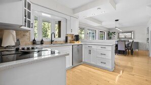 A modern kitchen with white cabinetry, a built-in stove, and a dining area with a table and chairs. - Capilano Forest Retreat | 3BD/2BA Suite (North Vancouver)