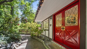A pathway leading to a red door with a stone base, surrounded by a garden with various plants and a wooden fence. - Capilano Forest Retreat | 3BD/2BA Suite (North Vancouver)