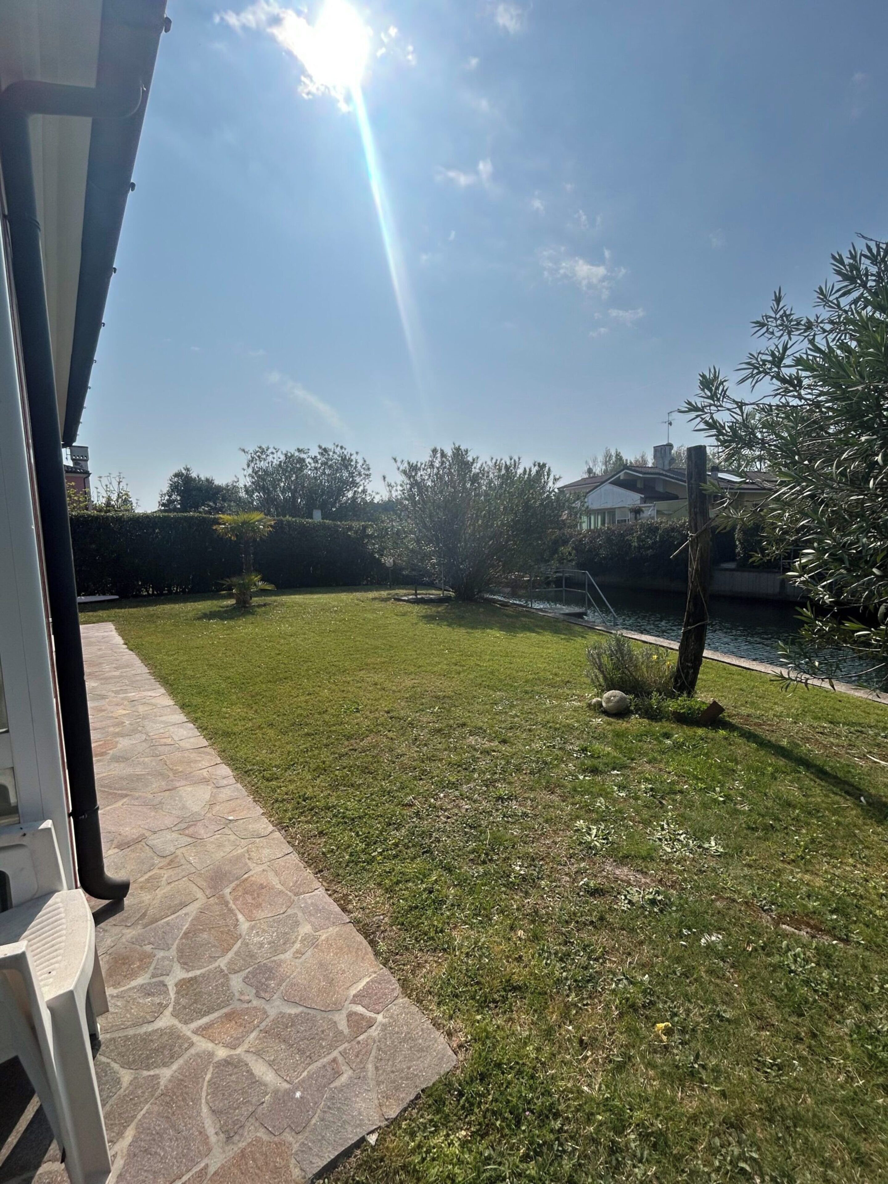 A grassy outdoor area with a stone pathway, a small tree, and a body of water.