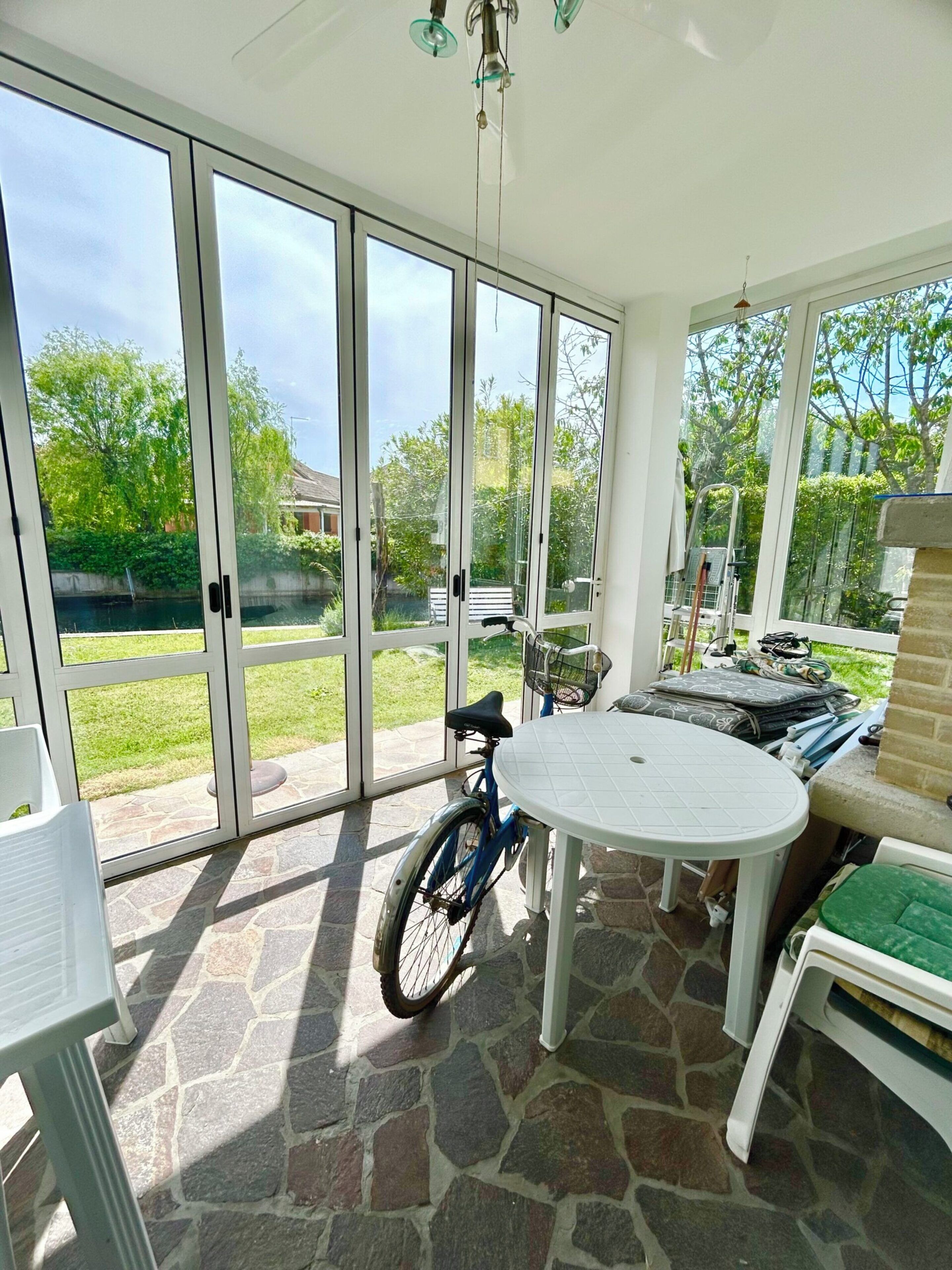 A covered patio with a stone floor, a table, chairs, a bicycle, and a view of a green garden through glass doors.