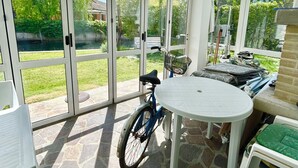 Um alpendre coberto com chão de pedra, uma mesa, cadeiras, uma bicicleta e vista para um jardim verde através de portas de vidro.