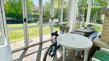 A covered patio with a stone floor, a table, chairs, a bicycle, and a view of a green garden through glass doors.