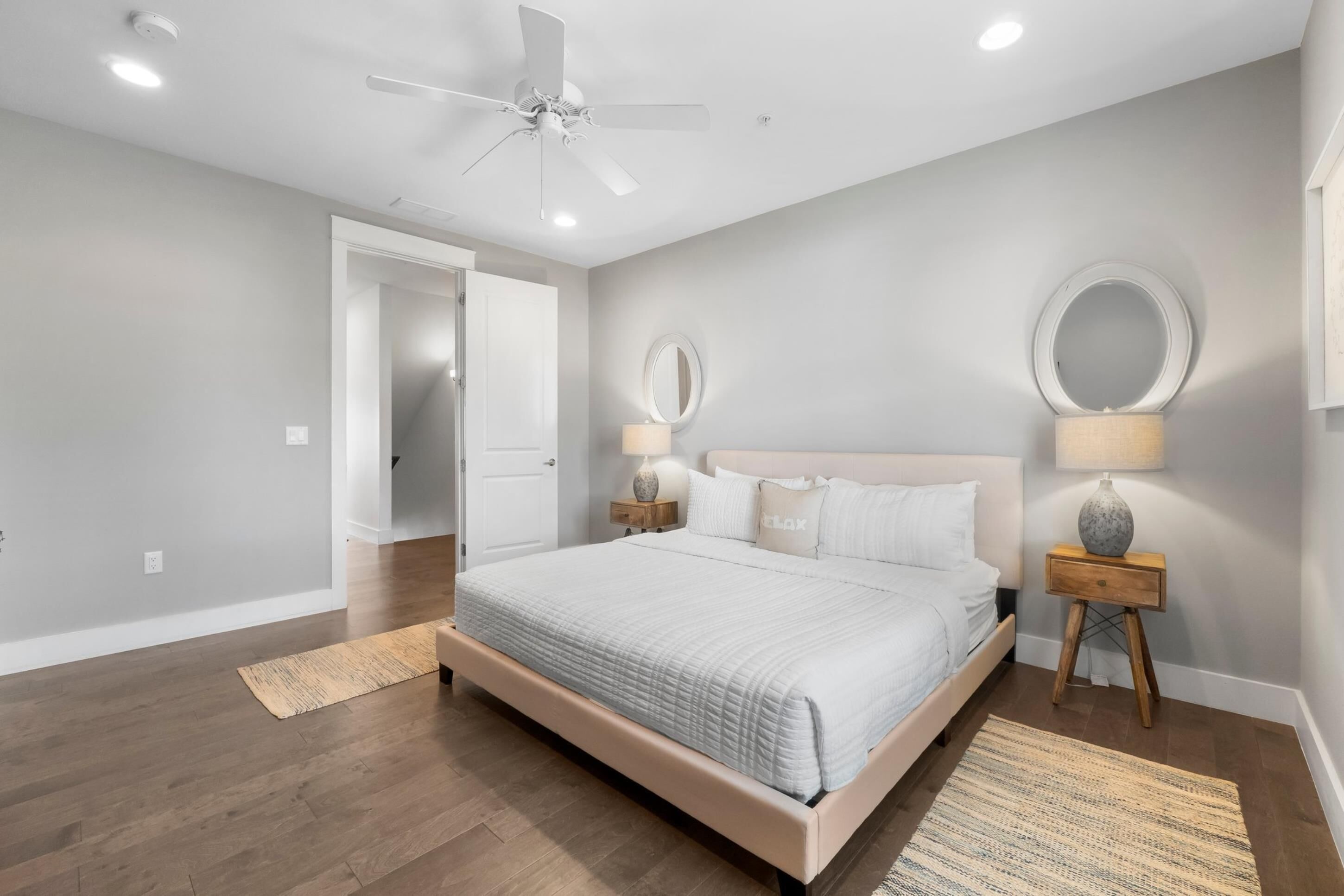 A bedroom with a bed, two bedside tables with lamps, a ceiling fan, and a door leading to another room.